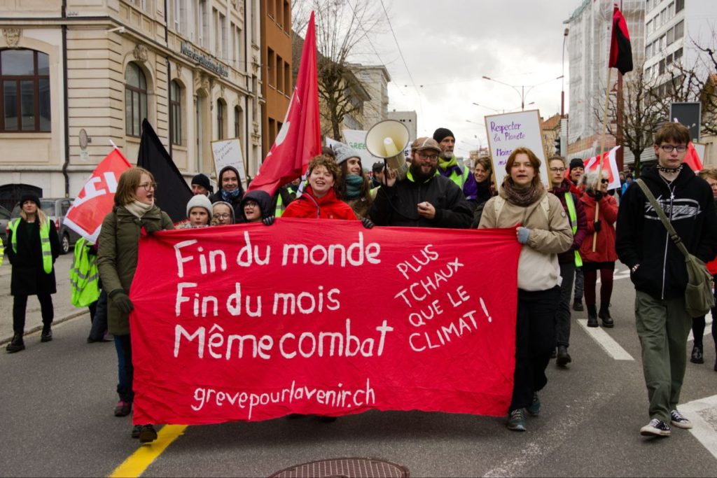 Photo de manifestantexs, banderole de tête sur laquelle il est écrit "fin du monde, fin du mois, même combat", "plus Tchaux que le climat" et "grevepourlavenir.ch"
